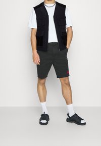 Mężczyzna w białej koszulce, czarnej kamizelce roboczej z kieszeniami, ciemnych spodniach cargo, białych skarpetkach i czarnych sandałach stoi na białym tle.