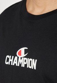 Sort bomulds-t-shirt med et hvidt "CHAMPION" logo med en rød accent. U-halsen er rund og ribbet, hvilket giver en enkel stil.
