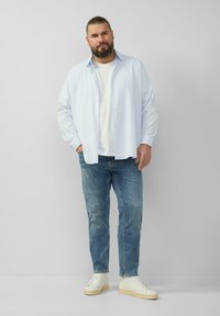 Lichtblauw overhemd over een wit t-shirt, gecombineerd met blauwe jeans en witte sneakers. Eenvoudige achtergrond, casual styling.