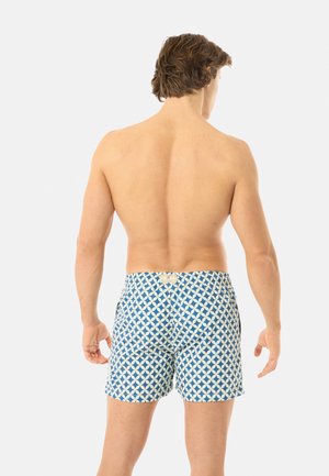Homme debout, vu de dos, portant un short de bain à motifs géométriques bleus et blancs, sur un fond uni.