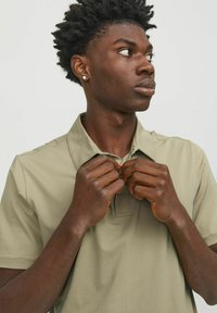 Polo shirt verde chiaro, maniche corte, colletto, materiale in cotone. Persona che aggiusta il colletto, con capelli scuri e texturizzati e sfondo neutro.