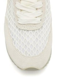 Zapatilla blanca con parte superior de malla con un patrón de panal, acentos de ante en un beige claro y cordones planos. Diseño de suela curvada.
