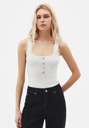 Femme blonde portant un haut blanc sans manches côtelé avec quatre boutons dorés et un jean noir taille haute sur un fond uni.