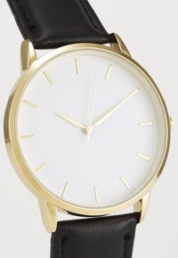Montre dorée avec un cadran blanc, des aiguilles slim pour les heures et les minutes, et un bracelet en cuir noir. Présente des marqueurs dorés minimalistes pour les heures.