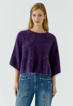 Femme aux longs cheveux blonds portant un pull violet ample et duveteux et un jean bleu taille haute avec de grandes poches avant sur un fond uni.
