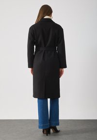 Cappotto di lana nero con vita alta e vestibilità rilassata, dotato di un colletto alto. Abbinato a jeans blu a campana e stivali neri con tacco alto.