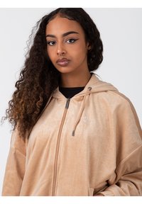 Sudadera con cremallera en color beige, con un tejido texturizado, capucha y cordones ajustables. Incluye un logo sutil en el pecho y un corte relajado.