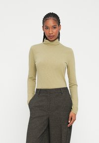 Mouwloze coltrui in licht olijfgroen, gemaakt van zacht materiaal. Gecombineerd met geruite, hoog-geplaatste broek in donkere tinten.