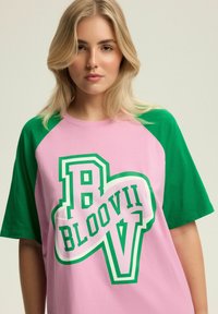 Młoda kobieta z blond włosami nosi różowo-zieloną koszulkę z napisem "B BLOOVII V" nadrukowanym dużymi literami z przodu.
