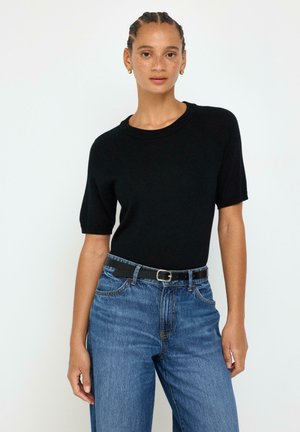 Donna che indossa una maglietta nera a maniche corte infilata in jeans blu con cintura nera, in piedi davanti a uno sfondo chiaro e semplice.