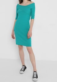 Türkisfarbiges Off-Shoulder-Kleid mit Dreiviertelärmeln, figurbetontem Schnitt und Überkreuz-Design. Kombiniert mit schwarz-weißen Sneakers.