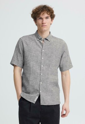 Jeune homme portant une chemise grise à manches courtes boutonnée et un pantalon noir, debout avec une main dans la poche devant un fond uni.