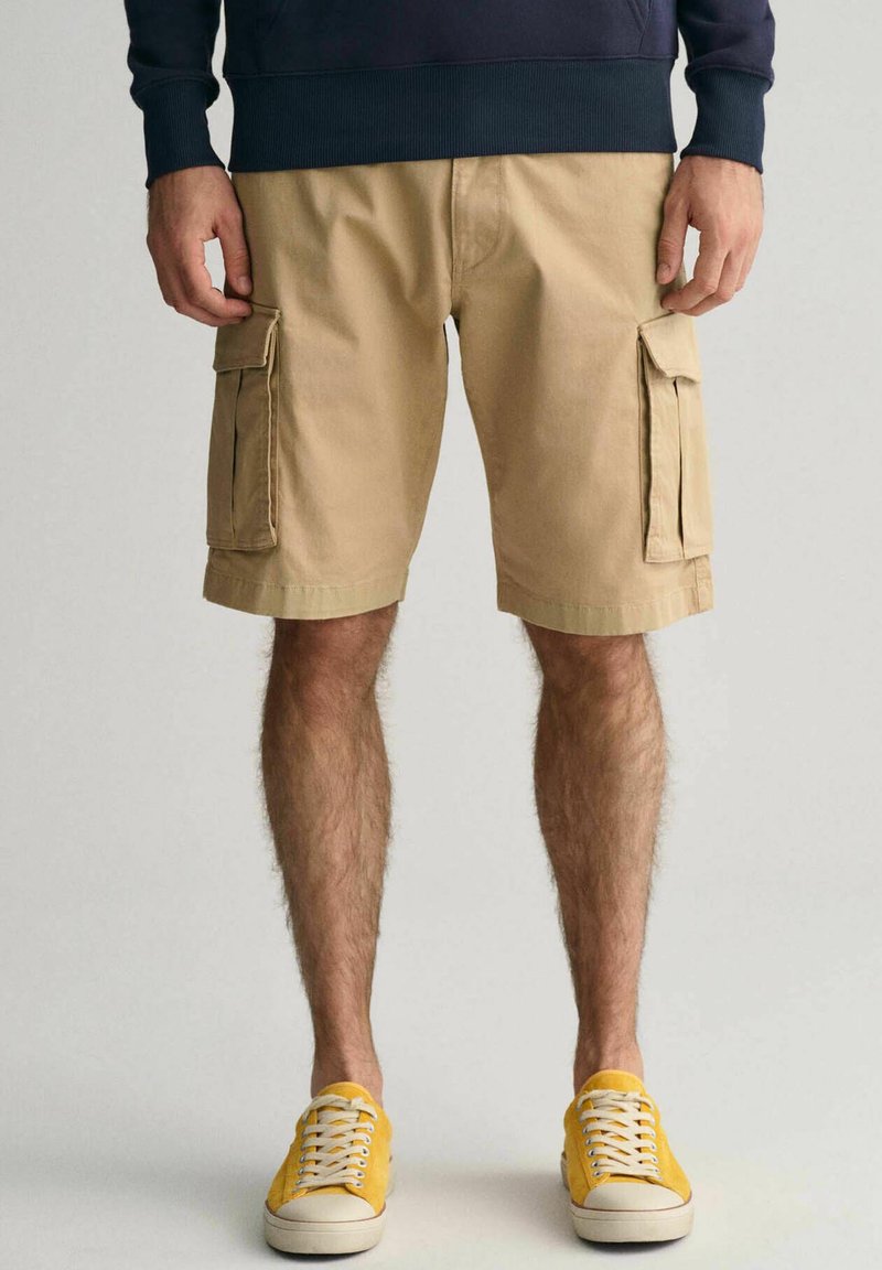 Beige cargo shorts med to sidelommer med klap og en løs pasform, parret med gule sneakers og en marineblå sweatshirt.