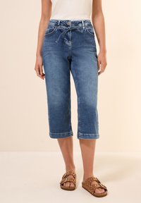 Keskisininen denim capri-housut, joissa on nauhavyötärö, etutaskut ja käännetyt lahkeet. Yhdistetty ruskeisiin sandaaleihin, joissa on niittejä.