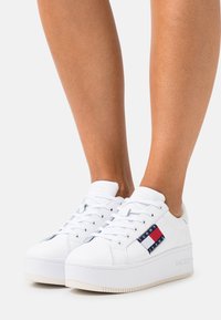 Bílé platformové tenisky z syntetického materiálu s logem v barvách červené, bílé a modré. Zavazování na tkaničky, kulatá špička.