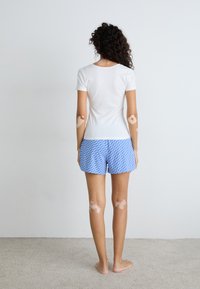 Vit t-shirt med kort ärm i kombination med blå mönstrade shorts som har ett upprepande mönster. Modellen står barfota mot en neutral bakgrund.