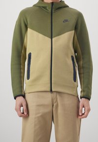 Hombre vestido con una sudadera con cremallera de Nike en colores oliva y beige, con cremallera negra y detalles en los bolsillos, combinada con pantalones beige, de pie contra un fondo liso.