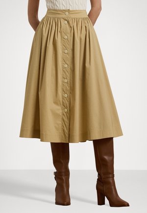 Kobieta w beżowej, midi spódnicy zapinanej na guziki, zestawionej z wysokimi brązowymi skórzanymi botkami na obcasie na białym tle.