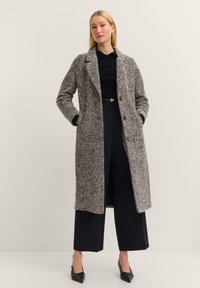 Langer grauer Hahnentrittmantel mit Reverskragen, Knopfverschluss und Taschen, kombiniert mit einem schwarzen gerippten Oberteil und weiten schwarzen Hosen.