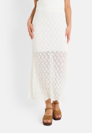 Jupe maxi en dentelle blanche avec un panneau inférieur transparent et un motif florale détaillé. Associée à des sandales à talons marron.