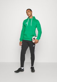 Haut à capuche vert avec cordons blancs et logo, pantalon noir avec des rayures blanches, tenant un ballon de soccer à motifs, portant des crampons noirs.
