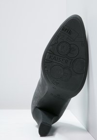 Chaussure à talon haut noire avec une surface lisse, présentant un talon structuré et une semelle embossée avec le nom de la marque "KAISER" et des détails décoratifs.