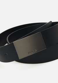 Sort læderbælte med en glat overflade, prydet med en rektangulær grå metalspænde præget med brandnavnet "HUGO."