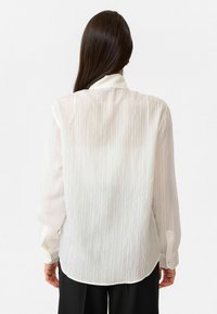 Witte gestreepte shirt gemaakt van lichtgewicht stof, met lange mouwen en een afgeronde zoom. De achterkant toont een zachte, doorzichtige textuur.