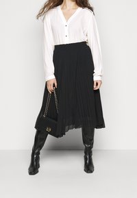 Donna in una blusa bianca e una gonna plissettata nera sta in piedi con sicurezza, tenendo una piccola borsa nera, mostrando un outfit elegante.