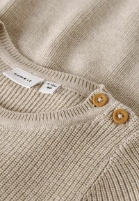 Maglione beige lavorato a maglia con scollatura a girocollo, caratterizzato da una texture a costine e due bottoni rotondi marroni sulla spalla. Etichetta della taglia visibile.