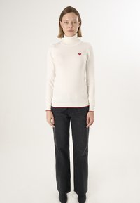 Crème kleurige turtleneck trui met een rood hartembleem, gecombineerd met donkere recht gesneden jeans; heeft rode accenten bij de manchetten en de zoom.