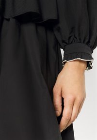 Svart klänning med en lager-på-lager, flödande design. Puffärmar med elastiska manschetter och en volangkant. Slät textur. Hand vilande nära fållen.