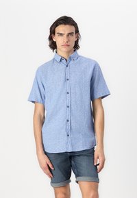 Chemise à manches courtes en tissu bleu clair avec un motif texturé, dotée d'un col et de boutons marron. Portée avec un short en denim.
