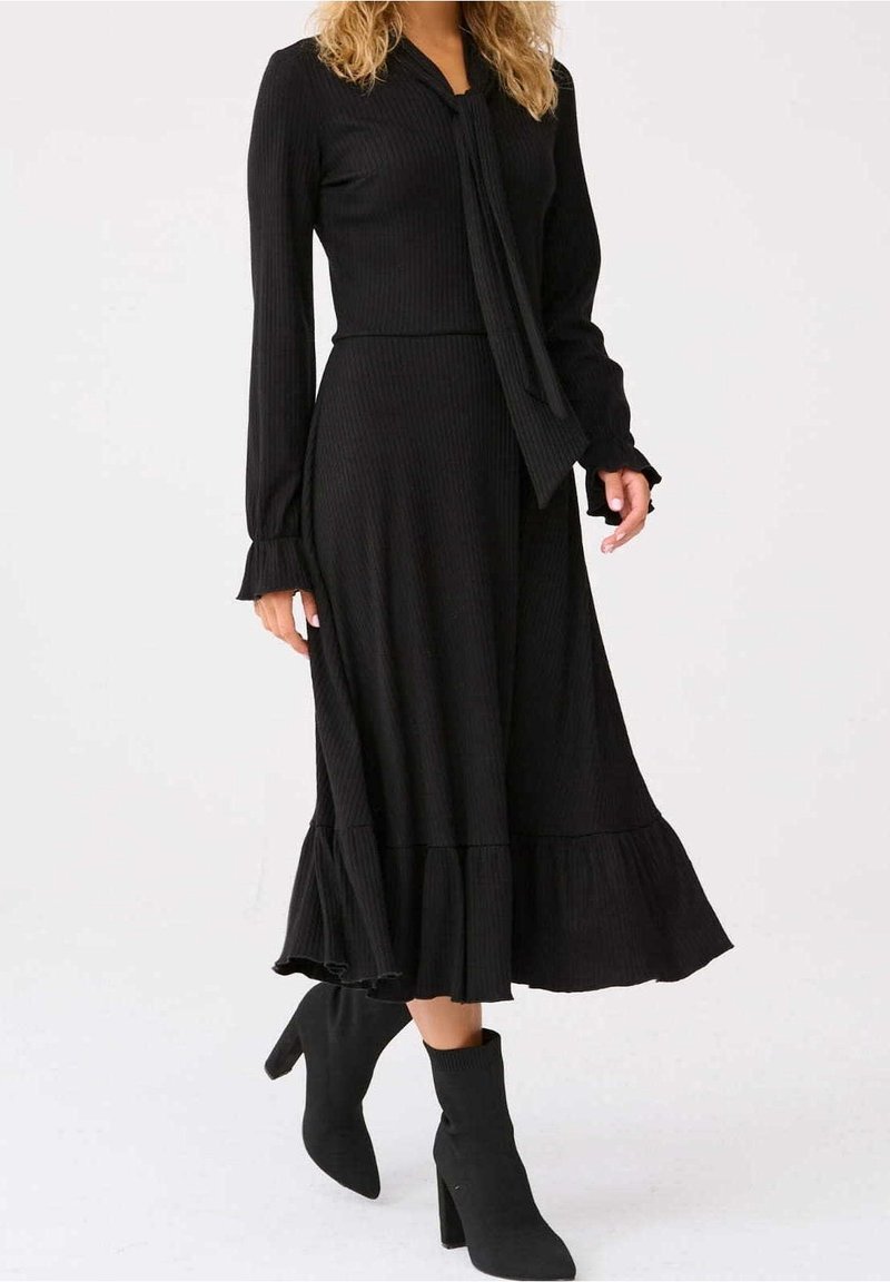 Femme portant une robe midi noire à manches longues avec des volants aux poignets et à l'ourlet, associée à des bottines noires à talons.