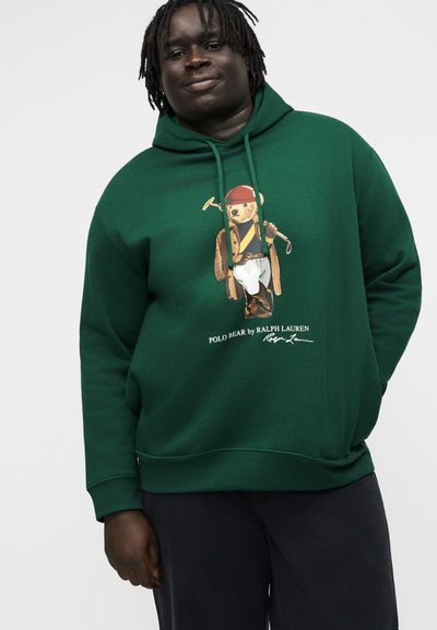 Oseba v temno zeleni kapucarji s potiskom Polo medveda v jahalni opremi na sprednji strani, združena s temnimi hlačami.