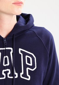 Sudadera con cremallera de color azul marino, hecha de un tejido suave, con un gran logotipo bordado en blanco "GAP" en el frente y una capucha forrada.