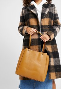 Torebka tote w odcieniu brązowym z fakturą, z złotymi detalami, o sztywnej konstrukcji i podwójnymi uchwytami, zestawiona z płaszczem w kratę.