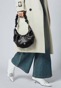 Sac à main noir et brillant avec un design décontracté et une anse froncée, associé à un trench-coat clair et un pantalon en denim à jambes larges.