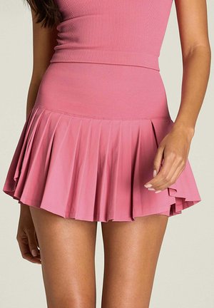 Vrouw met een roze geribd topje en bijpassende roze geplooide minirok, staand met één hand langs haar zijde.