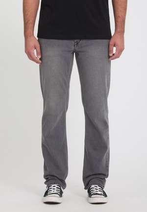 Homme portant un jean gris, un t-shirt noir et des baskets noires avec des lacets blancs, debout devant un fond uni.