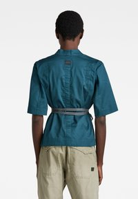Personne aux cheveux courts et bouclés porte une chemise à manches courtes teal avec une fine ceinture nouée à la taille et un pantalon beige, vue de dos.