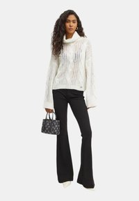 Maglione bianco lavorato a maglia con colletto a dolcevita e design a treccia, abbinato a pantaloni neri a zampa e una piccola borsa nera.