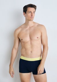 Boxer-briefs navy avec une ceinture jaune clair étiquetée "LACOSTE". Le tissu semble doux et extensible, conçu pour le confort et un bon ajustement.