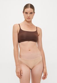 Vrouw met slicked-back haar, gekleed in een bruine crop top en beige hoge onderbroek tegen een vlakke witte achtergrond.
