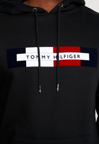 Svart hoodie med en central logotyp i rött, vitt och marinblått. Har dragsnören med metallspetsar och en mjuk textur.