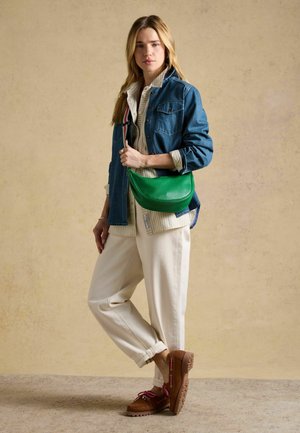 Kvinde iført blå denimjakke, stribet skjorte, cremefarvede bukser, brune mokkasiner, bærende en grøn crossbody-taske mod en beige baggrund.