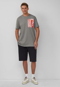 Graues T-Shirt mit kurzen Ärmeln und einem bunten Grafik-Taschen-Design. Kombiniert mit dunkelgrauen Shorts und weißen Sneakers. Schlichte, lässige Passform.