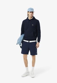 Felpa navy con tasca a marsupio, pantaloni corti navy con logo, giacca di jeans sulle spalle, cappellino azzurro chiaro e sneakers bianche.