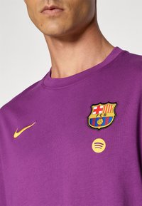 Muškarac nosi ljubičastu sportsku majicu s žutim Nike swooshom, grbom FC Barcelone i žutim Spotify logotipom na prsima.