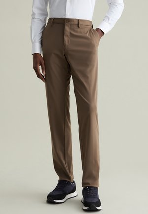 Pantalon brun avec une texture lisse, coupe droite et poches latérales, associé à des baskets sombres et une chemise blanche à manches longues.